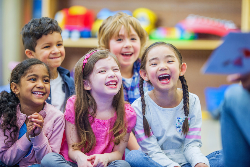 A diverse group of preschoolers inside the classroom sitting on the floor laughing and listening to their teacher reading a book