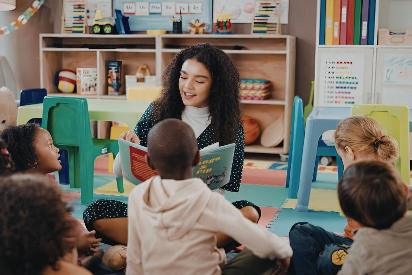 Childcare teacher sits on the floor with a group of children and reads a book to them