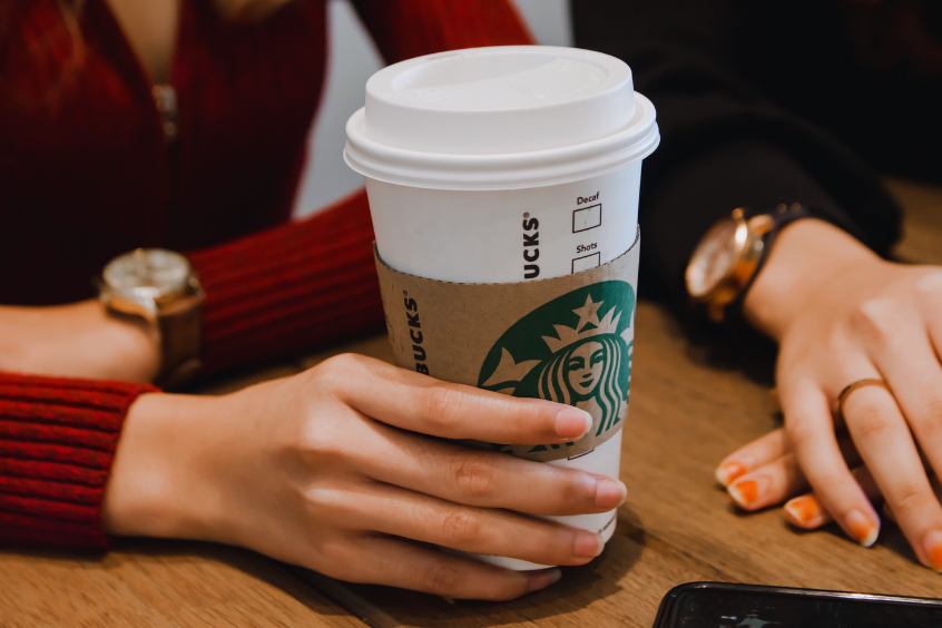 A person holding a Starbucks takeaway coffee cup at a café table